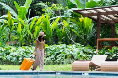 Havuz kenarında turuncu bavullu, şapkalı ve şık giyinmiş bir kadın. Arkaplanda verimli bir yeşillik ve ahşap bir yapı var. Tropik bir tatil ortamı yaratıyor..