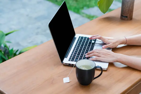 Tahta bir masanın üzerine dizüstü bilgisayarda yazan biri. Laptopun yanında bir fincan çay ya da kahve var. Arka planda yeşillik var gibi görünüyor.