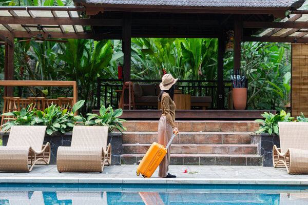 A woman walking by a poolside with an orange suitcase, wearing a hat and stylish outfit. The background features lush greenery and a wooden structure, creating a tropical vacation vibe.