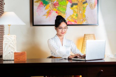 Beyaz bluzlu bir kadın, duvarında renkli bir resimle iyi aydınlatılmış bir odada dizüstü bilgisayarda çalışıyor..