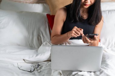 Yatakta oturan genç bir kadın, dizüstü bilgisayar kullanırken akıllı telefonuna odaklanmış. Uzun siyah saçları var ve siyah bir bluz giyiyor. Yatak beyaz yatak ve kırmızı yastıkla düzgünce yapılmıştır.