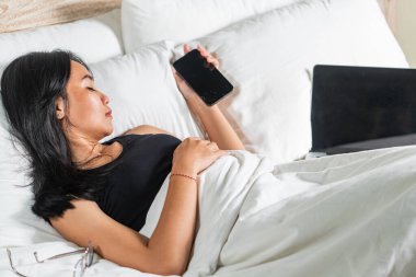 Battaniyeye sarılmış, akıllı telefonuna bakan genç bir kadın. Yatağın üzerine bir dizüstü bilgisayar yerleştiriliyor. Ortam rahat ve rahat, arka planda yumuşak yastıklar var..