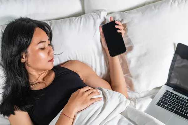 Battaniyeye sarılmış, akıllı telefonuna bakan genç bir kadın. Yatağın üzerine bir dizüstü bilgisayar yerleştiriliyor. Ortam rahat ve rahat, arka planda yumuşak yastıklar var..