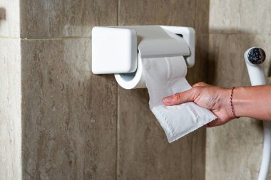 Tuvalet kağıdına uzanan bir el. Arka planda duvarlar ve tuvalet var. Odak noktası el ve tuvalet kağıdı rulosu..