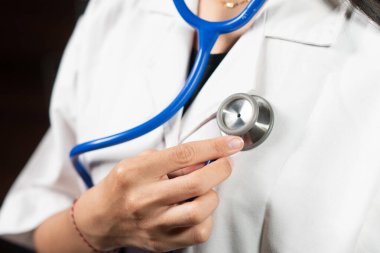 Göğsüne steteskop dayamış beyaz laboratuvar önlüğü giyen bir sağlık uzmanının yakın çekimi. Steteskop mavidir ve kişi klinik ortamdadır..