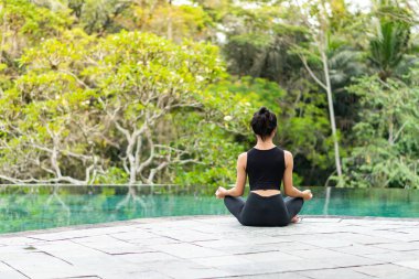 Serin bir sonsuzluk havuzunun kenarında oturan bir kadın, yemyeşil ile çevrili. Meditasyon pozisyonunda, doğanın sakin atmosferinin tadını çıkarıyor. Sahne huzur ve rahatlık verir..