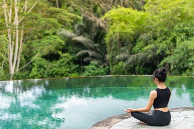 Serin bir sonsuzluk havuzunun kenarında oturan bir kadın, yemyeşil ile çevrili. Meditasyon pozisyonunda, doğanın sakin atmosferinin tadını çıkarıyor. Sahne huzur ve rahatlık verir..