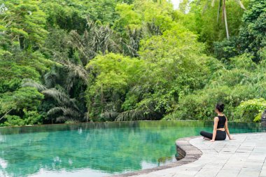 Serin bir sonsuzluk havuzunun kenarında oturan bir kadın, yemyeşil ile çevrili. Meditasyon pozisyonunda, doğanın sakin atmosferinin tadını çıkarıyor. Sahne huzur ve rahatlık verir..