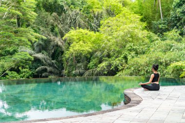 Serin bir sonsuzluk havuzunun kenarında oturan bir kadın, yemyeşil ile çevrili. Meditasyon pozisyonunda, doğanın sakin atmosferinin tadını çıkarıyor. Sahne huzur ve rahatlık verir..