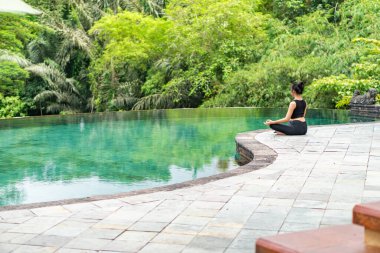 Serin bir sonsuzluk havuzunun kenarında oturan bir kadın, yemyeşil ile çevrili. Meditasyon pozisyonunda, doğanın sakin atmosferinin tadını çıkarıyor. Sahne huzur ve rahatlık verir..