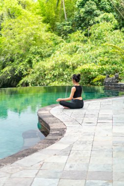 Serin bir sonsuzluk havuzunun kenarında oturan bir kadın, yemyeşil ile çevrili. Meditasyon pozisyonunda, doğanın sakin atmosferinin tadını çıkarıyor. Sahne huzur ve rahatlık verir..