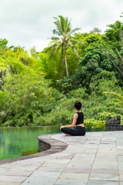 Serin bir sonsuzluk havuzunun kenarında oturan bir kadın, yemyeşil ile çevrili. Meditasyon pozisyonunda, doğanın sakin atmosferinin tadını çıkarıyor. Sahne huzur ve rahatlık verir..