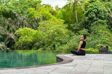 Serin bir sonsuzluk havuzunun kenarında oturan bir kadın, yemyeşil ile çevrili. Meditasyon pozisyonunda, doğanın sakin atmosferinin tadını çıkarıyor. Sahne huzur ve rahatlık verir..