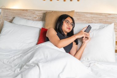 Genç bir kadın akıllı telefon kullanarak yatakta yatıyor. Beyaz bir battaniyeyle örtülmüş ve yatak odası sıcacık yastıklarla çevrili. Işık yumuşak, rahat bir atmosfer yaratıyor..