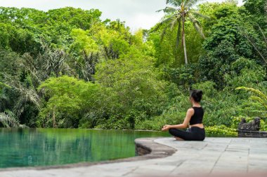 Serin bir sonsuzluk havuzunun kenarında oturan bir kadın, yemyeşil ile çevrili. Meditasyon pozisyonunda, doğanın sakin atmosferinin tadını çıkarıyor. Sahne huzur ve rahatlık verir..