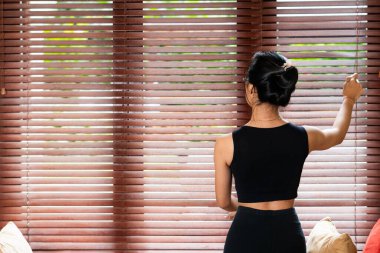 Siyah kolsuz tişörtlü bir kadın rahat bir odada ahşap panjurları ayarlıyor. Sırtı kameraya dönük duruyor, panjurlara odaklanıyor. Yumuşak doğal ışık tabakaların arasından süzülür.