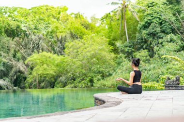 Serin bir sonsuzluk havuzunun kenarında oturan bir kadın, yemyeşil ile çevrili. Meditasyon pozisyonunda, doğanın sakin atmosferinin tadını çıkarıyor. Sahne huzur ve rahatlık verir..