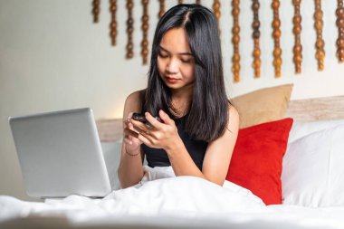 Yatakta oturan genç bir kadın, dizüstü bilgisayar kullanırken akıllı telefonuna odaklanmış. Uzun siyah saçları var ve siyah bir bluz giyiyor. Yatak beyaz yatak ve kırmızı yastıkla düzgünce yapılmıştır.