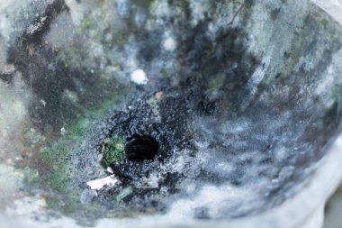 Merkezinde karanlık, dairesel bir delik olan yıpranmış taş bir leğenin yakın çekim görüntüsü. Yüzeyde yosun ve kir birikimi izleri var. Bu da yaşı ve ihmali gösteriyor..