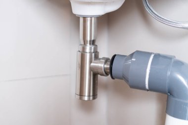 Lavabonun altındaki tesisat borularına yakın plan, paslanmaz çelik konnektör ve gri plastik boru var. Resim, farklı boru materyalleri arasındaki bağlantıyı vurgular.