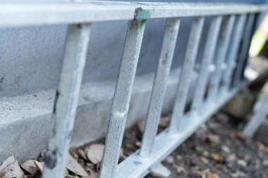 Duvara dayanmış, basamaklara ve dokulara odaklanmış metalik bir merdiven görüntüsü. Toprak küçük taşlar ve molozlarla kaplı, kırsal bir ortam yaratıyor..