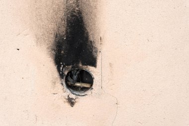 A close-up view of a round hole in a beige wall, showing signs of smoke damage around the edges. The interior of the hole reveals some debris, possibly sticks or wires, indicating a potential entry point or vent. clipart