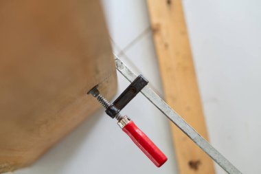 Ahşap işçiliğinin kırmızı saplı klempine yakın çekim, ahşap bir parçayı emniyete alıyor. Arkaplanda, atölye ortamını vurgulayan ahşap kirişler yer alıyor.