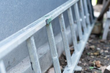 Duvara dayanmış metal bir merdiveni yakından görmek. Yerdeki toprak ve döküntülerle kısmen gizlenmiş..