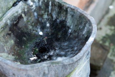 Karanlık lekeler ve yosunlarla kaplanmış yıpranmış taş bir leğenin yakından görüntüsü, içine akan küçük bir su akışını gösteriyor. Yüzey desenli ve yaşlıdır. Bu da elementlere uzun süreli maruz kaldığını gösterir..