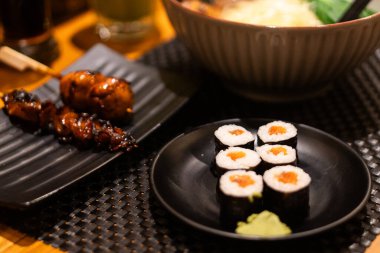 Somon dolgulu suşi dürümleri, yemek çubuklarıyla siyah bir tabakta tutuluyor. Arka planda bir kase çorba ve şiş et var. Japon mutfağı hazırlıyorlar..