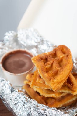 Alüminyum folyoya sarılı küçük bir fincan çikolata sosuyla servis edilen bir yığın altın kahverengi waffle..