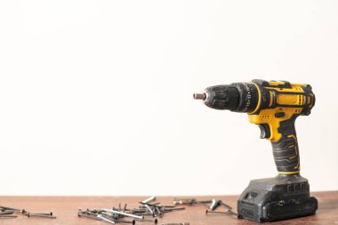 Etrafı dağınık vidayla çevrili ahşap bir yüzeye yerleştirilmiş sarı ve siyah bir kablosuz matkap. Arkaplan sade ve açık renkli, araçları vurguluyor.