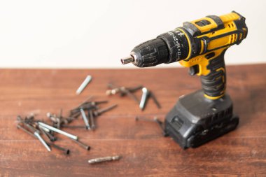 Tahta bir yüzeye yerleştirilmiş, çeşitli vida ve cıvatalarla çevrili sarı ve siyah bir kablosuz matkap. Matkap, tasarımını ve özelliklerini göstererek izleyiciye doğru açılıdır..