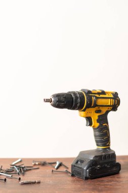 Tahta bir yüzeye yerleştirilmiş, etrafa saçılmış vida ve cıvatalarla çevrili sarı ve siyah kablosuz bir matkap. Arkaplan düz ve açık renkli, matkap ve donanımı vurguluyor.