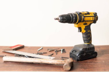 Tahta bir yüzeyde çekiç, vida ve kare gibi çeşitli aletlerle çevrili kablosuz bir matkap. Matkap sarı ve siyahtır, tasarımı ve işlevselliğini gösterir..