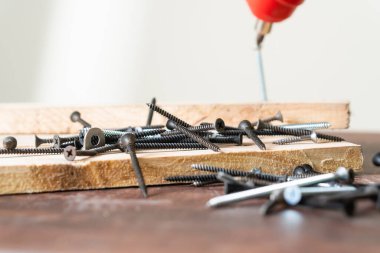 Ahşap bir yüzeye saçılmış çeşitli vidaların yakın plan görüntüsü, arka planda matkapla. Görüntü, tahtanın dokusunu ve vidaların metalik kısmını yakalar..