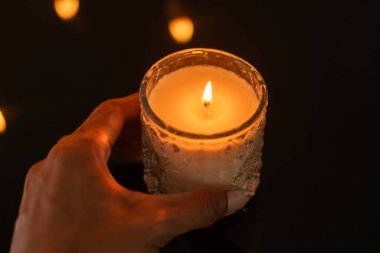 A hand holding a lit candle in a glass holder, with a dark reflective surface and soft glowing lights in the background. clipart