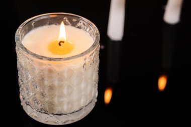 Kalıplı bir cam tutacağında, sıcak bir parıltıyla yanan bir mum. Arka planda, iki tane yanmamış mum görülebiliyor, sakin bir atmosfer yaratıyor..