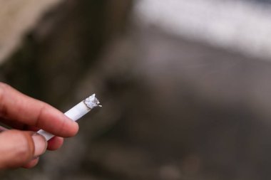 Ucundan duman yükselirken, yanan bir sigarayı tutan bir elin yakın çekimi. Arka plan bulanık, bu da sigaraya odaklanmamızı sağlıyor..