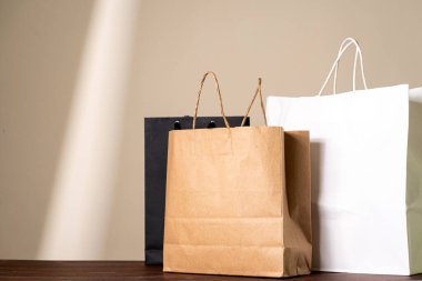 Üç alışveriş torbası farklı renklerde (kahverengi, siyah ve beyaz) bej arka planda, yumuşak ışıklandırmalı ahşap bir masaya yerleştirilmiştir..