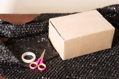 Pembe makas ve temiz bant eşliğinde siyah bir baloncuk ambalaj yüzeyine yerleştirilmiş kahverengi bir karton kutu. Ayarlar ambalaj veya nakliye faaliyetlerini öneriyor.