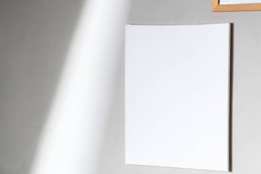 Açık gri bir duvara monte edilmiş boş beyaz bir tuval, yakınlardaki bir pencereden gelen yumuşak doğal ışıkla aydınlatılmış, sakin ve minimalist bir atmosfer yaratıyor..