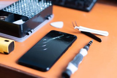 Çatlak ekranlı akıllı bir telefon tamir aletleriyle çevrili bir tezgahta yatar. Tornavida seti, plastik kazma ve cımbız da dahil..