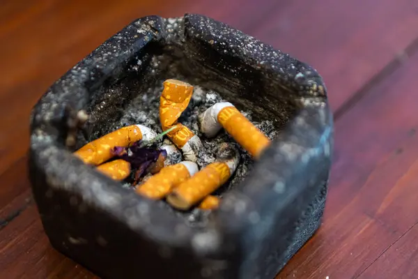 Kullanılmış sigara izmaritleri ve küllerle dolu bir kül tablası, ahşap bir yüzeyde duruyor. Kül tablası koyu ve desenli, yanmış tütün kalıntıları ve kurumuş çiçeklerle dolu..