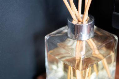 Ahşap kamışlı şık bir cam dağıtıcısı bir masada, yanan bir mumun yanında oturur. Arka planda bir bilgisayar klavyesi ve yumuşak ışıklı bir ekran var, rahat bir atmosfer yaratıyor..