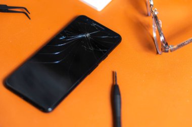 Turuncu bir masaya yerleştirilmiş kırık ekranlı akıllı bir telefon. Çevreleyen araçlar arasında cımbız, tornavida ve bir klavye ile bir çift gözlük ile birlikte, bir onarım veya teknik çalışma alanı öneren.