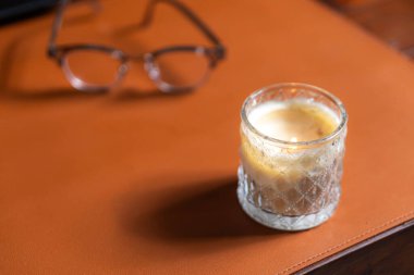 A lit candle in a glass holder sits on a brown leather surface, with a pair of eyeglasses in the background, creating a cozy atmosphere. clipart