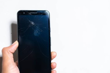 Beyaz arka planda kırık ekranlı akıllı bir telefonu tutan bir elin yakın çekimi. Ekran karanlık, örümcek ağı gibi çatlakları vurguluyor..