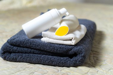 Beyaz bir pompa şişesi ve düzgünce katlanmış havlulara yerleştirilmiş sarı kaplı bir kap. Arka planda yumuşak, dokulu bir kumaş var, sakin bir atmosfer yaratıyor..