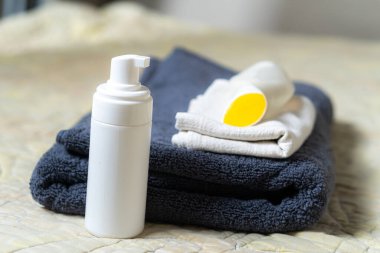 Beyaz bir pompa şişesi ve düzgünce katlanmış havlulara yerleştirilmiş sarı kaplı bir kap. Arka planda yumuşak, dokulu bir kumaş var, sakin bir atmosfer yaratıyor..